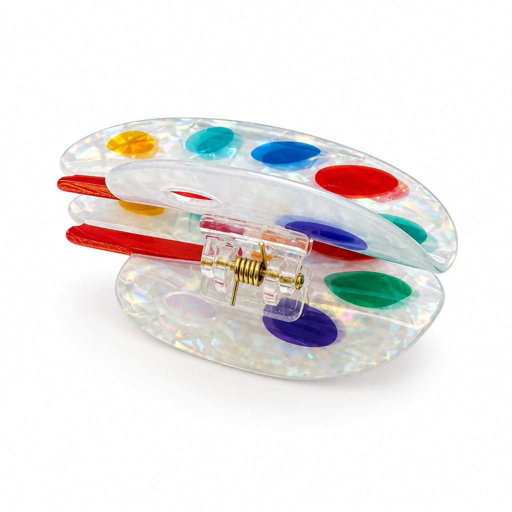 A clear plastic hair claw clip decorated with colorful paint splatters in red, blue, green, yellow, and purple, with red paintbrushes visible inside, set against a plain white background.